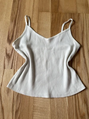 Vit ribbad linnetopp med smala band - Snygg vit ribbad linnetopp med smala axelband och v-ringad hals. Toppen är ärmlös och har en enkel, stilren design som passar perfekt till jeans eller kjol. Materialet är mjukt och stretchigt för en bekväm passform.