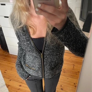 Grå stickad kofta med dragkedja - Fin kofta/ tunn jacka. Stickat tyg i svart, vitt och grått. Krage i skinn material. Koftan är köpt second hand och har lite slitningar i kragen och på skinn detaljerna. Två fickor framtill och guld kedja.