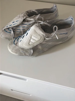 Adidas Predator vita fotbollsskor - Säljer ett par vita Adidas Predator fotbollsskor med blå detaljer och klassisk tunga över snörningen. Skorna har en modern, strömlinjeformad design och är tillverkade i syntetmaterial för bästa känsla på planen. Sula med dobbar och stor PREDATOR-text undertill.