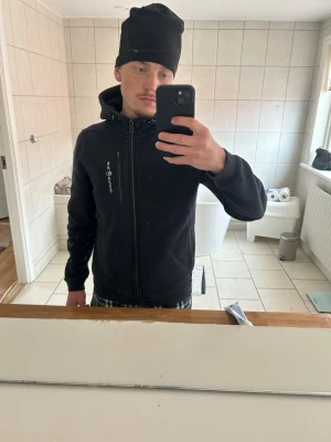Svart hoodie från Sail Racing - Svart hoodie från Sail Racing med dragkedja hela vägen, huva med dragsko och en bröstficka med dragkedja. Snygg logga på bröstet och mjukt material som känns skönt mot huden. Perfekt för en avslappnad och sportig stil.