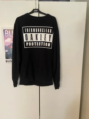 Svart Oakley sweatshirt med tryck - Cool svart sweatshirt från Oakley med stort vitt tryck på bröstet där det står 'THERMONUCLEAR OAKLEY PROTECTION'. Tröjan har rund hals, långa ärmar och ribbade muddar. Perfekt för en avslappnad streetwear-look. Skickar inom 24 timmar!