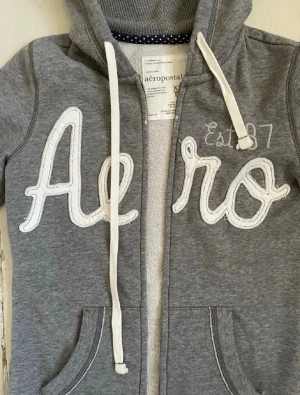 Grå Aeropostale hoodie med dragkedja - Grå hoodie från Aeropostale med vit broderad text 'Aero' och 'Est. 87' på bröstet. Tröjan har huva med vita snören, dragkedja framtill och två fickor. Mjuk insida och klassisk streetstil, perfekt för chill dagar. Skriv vid frågor💕