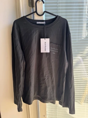 Svart långärmad topp Acne Studios - Svart långärmad topp från Acne Studios med rund halsringning och bröstficka med vit text 'Acne Studios STOCKHOLM 1996'. Tillverkad i mjuk bomull, perfekt för en clean och stilren look.