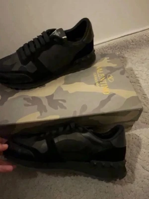 Valentino svarta camo sneakers - Snygga svarta sneakers från Valentino med camo-mönster och detaljer i mocka och textil. Skorna har en låg profil, svarta snören och en robust sula med nitar baktill. Perfekta för dig som gillar exklusiva och trendiga sneakers.