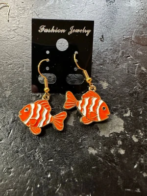 Färgglada fiskörhängen i emalj - Snygga örhängen i form av orange och vita fiskar med svarta detaljer. Hängena är i emalj med guldfärgade krokar. Perfekt accessoar för dig som gillar unika och lekfulla smycken. 