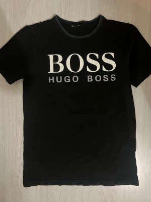 Svart Hugo Boss t-shirt med tryck - Svart t-shirt från Hugo Boss med stort vitt logotryck på bröstet. Klassisk rund halsringning och korta ärmar. Perfekt för en stilren och enkel look.