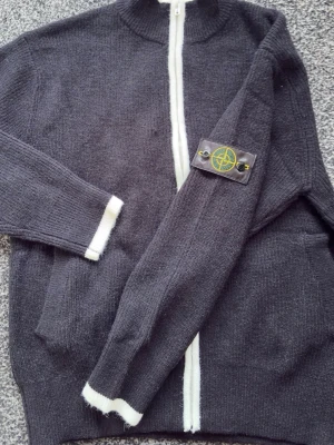 Stone Island grå stickad kofta - Mörkgrå stickad kofta från Stone Island med vit dragkedja och vita detaljer vid ärmslut och nederkant. Klassisk patch på ena ärmen och två fickor framtill. Perfekt för lager-på-lager och snygg till jeans.