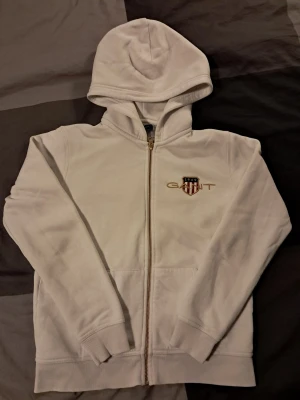 Vit hoodie med dragkedja från Gant - Snygg vit hoodie från Gant med dragkedja och broderad logga på bröstet. Klassisk design med huva och kängurufickor framtill. Mjuk insida för extra komfort, perfekt för en avslappnad stil.
