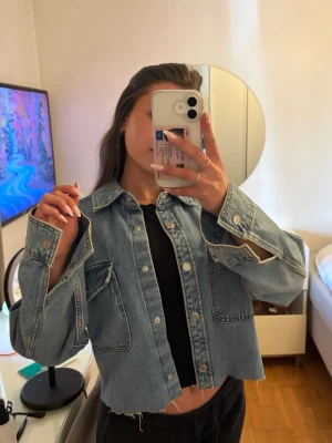 Cropped blå jeansjacka - Säljer en trendig cropped jeansjacka i klassisk blå denim med rå kant nertill. Jackan har stora bröstfickor med lock, silverfärgade knappar och breda ärmslut. Perfekt för dig som vill ha en avslappnad och cool stil.