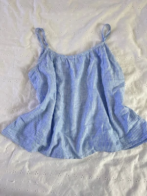 Blåvit randig linnetopp från H&M - Säljer en söt blåvit randig linnetopp från H&M med tunna axelband och avslappnad passform. Perfekt för varma dagar och enkel att styla med jeans eller kjol. Luftigt material som känns skönt mot huden.