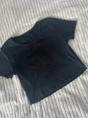 Svart ribbad croppad topp - En svart croppad topp med ribbad struktur och korta ärmar. Toppen har rund halsringning och är tillverkad i ett stretchigt material som sitter snyggt på kroppen. Perfekt att styla med jeans eller kjol för en trendig look.