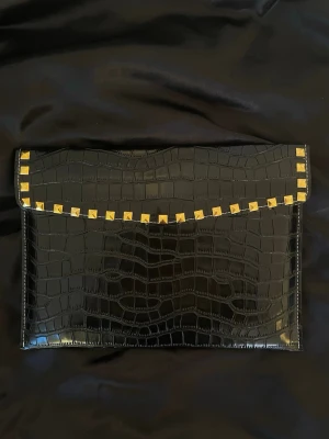 Datorfodral i krokodil mönster med guldnitar  - Stilrent datorfodral tillverkat av veganskt läder i vattentätt material. Fodralet stängs med knapp. Fodralet skyddar din dator effektivt samtidigt som det ger din outfit en extra touch. ⚜️ Storlek: 34 × 25 cm –  Innifrån 33 x 24 📦 