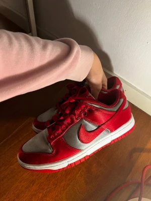 Nike Dunk Low röd och grå - Nike Dunk Low sneakers i rött och grått med klassisk Swoosh-logga på sidan. Skorna har röd ovandel i läder och grå paneler i textil, samt röda skosnören och vit mellansula med röd yttersula. Perfekt för dig som gillar streetwear och vill sticka ut.