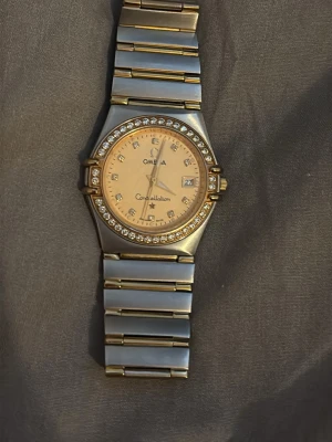 Omega Constellation klocka silver/guld - Snygg Omega Constellation klocka i silver och guld med länkarmband i metall. Urtavlan är beige med datumvisning och omgiven av glittrande stenar. Klassisk och exklusiv design med tydliga index och gulddetaljer.