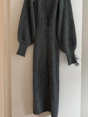 Stickad klänning Adoore - Lång, ribbstickad kofta/klänning i mörkgrått från Adoore. Koftan har ballongärmar, v-ringning och knappar längs hela framsidan. Tillverkad i ett mjukt, tjockt material som ger en mysig känsla. Perfekt för lager på lager och chill dagar.