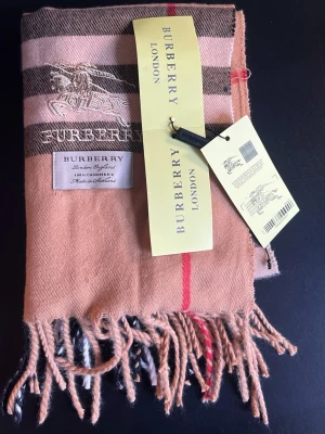 Beige rutig halsduk från Burberry - Klassisk beige halsduk från Burberry i 100% kashmir, tillverkad i Skottland. Halsduken har det ikoniska rutmönstret i beige, brunt, vitt och rött samt fransar i ändarna. Broderad Burberry-logga och etikett framtill. Perfekt accessoar för en stilren look.