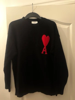 Svart stickad tröja från Ami Paris - Svart stickad tröja från Ami Paris med röd hjärtlogga och bokstaven A på bröstet. Tröjan har rund halsringning och långa ärmar. Perfekt för dig som gillar stilrena plagg med en twist.