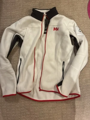 Vit fleecejacka från Helly Hansen - Vit fleecejacka från Helly Hansen med röda detaljer och svarta inslag. Jackan har dragkedja framtill, två fickor med dragkedja och hög krage. Perfekt för kyliga dagar och outdoor-aktiviteter. Skulle inte säga att den sitter så löst utan taigjt. Känns som M