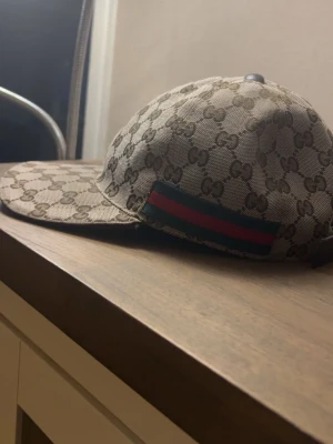 Gucci beige GG keps med röd-grön rand - Snygg beige keps från Gucci med klassiskt GG-monogram över hela kepsen och en ikonisk röd-grön rand på sidan. Kepsen har böjd skärm och justerbar rem bak. Tillverkad i canvasmaterial med svart insida. Perfekt för dig som vill sticka ut med lyxig streetstyle.