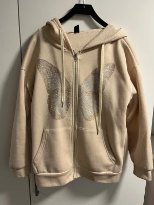 Beige hoodie med fjärilstryck - Snygg beige hoodie med dragkedja och stor huva. På framsidan finns ett stort glittrigt fjärilstryck i silver. Hoodien har två rymliga fickor framtill och ribbade muddar vid ärmar och nederkant. Perfekt för en chill och trendig look. Köpt på SHEIN och använd ett fåtal gånger💗