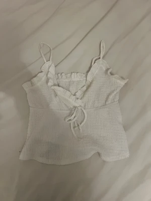 Vit linnetopp med volang och knyt - Söt vit linnetopp med smala axelband, volangkant upptill och dekorativ knytning framtill. !!Om du köper välj gärna att hämta upp på utlämningsställe då det blir enklare för mig att posta på det viset!!
