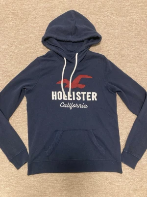 Mörkblå hoodie från Hollister - Mörkblå hoodie från Hollister med vit dragsko i huvan och stor logga i vitt och rött på bröstet. Klassisk känguruficka framtill och mjukt material som känns skönt mot huden. Perfekt för en avslappnad och stilren look.