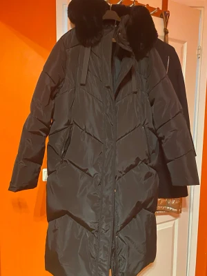 Svart lång dunjacka med fuskpäls - Snygg svart lång dunjacka med chevronmönster och stor huva med fuskpäls. Jackan har dragkedja framtill, två sidofickor med dragkedja och är perfekt för kalla vinterdagar. Oversized passform och stilren look.