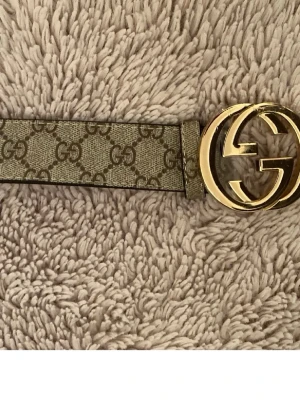Gucci GG bälte - Snyggt Gucci-bälte med klassiskt GG-mönster i beige och brunt. Bältet har ett stort, rundat spänne i guld med den ikoniska GG-loggan. Perfekt accessoar för att lyfta vilken outfit som helst.