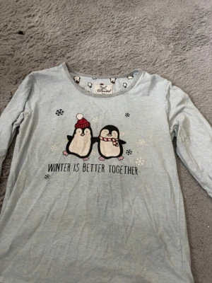 Ljusgrå pyjamas med pingvinmotiv - Supermysig ljusgrå pyjamas från Cool Club med långärmad tröja och byxor. Tröjan har gulligt pingvintryck och texten 'Winter is better together', medan byxorna har ett all-over pingvinmönster och dragsko i midjan. Perfekt för vinterkvällar och chill hemma.