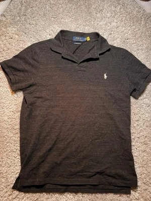 Mörkgrå pikétröja från Polo Ralph Lauren - Klassisk mörkgrå pikétröja från Polo Ralph Lauren med vit broderad logga på bröstet. Tröjan har korta ärmar, krage och knappar vid halsen. Nyskick knappt använd, storlek M. Pm för mer info