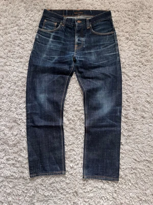 Mörkblå jeans från Nudie Jeans - Snygga mörkblå jeans från Nudie Jeans med raka ben och klassisk femficksdesign. Jeansen har tydliga slitningar och kontrastsömmar samt läderpatch bak i midjan. Tillverkade i kraftig denim, perfekt för en avslappnad stil.