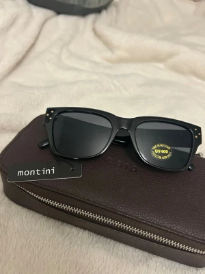 Svarta solglasögon från Montini - Stilrena svarta solglasögon från Montini med klassisk rektangulär form och mörka glas. Bågarna har små guldfärgade detaljer vid hörnen och ger ett coolt intryck. Perfekta för soliga dagar och skyddar med UV400-filter.