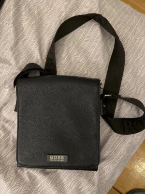 Svart axelväska från Hugo Boss - Snygg svart axelväska från Hugo Boss i stilren design. Väskan är tillverkad i strukturerat läder med justerbar axelrem i textil och metallogo framtill. Perfekt storlek för det viktigaste och passar till många outfits.