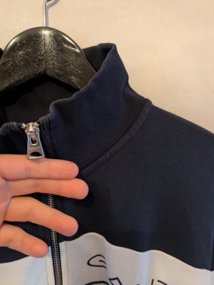 Marinblå zip hoodie från GANT - Marinblå hoodie från GANT med vit och blå randig detalj över bröstet och logga på vänster sida. Dragkedja framtill och ribbade muddar vid ärmar och midja. Tillverkad i mjukt bomullsmaterial och har en hög krage för extra stilpoäng.