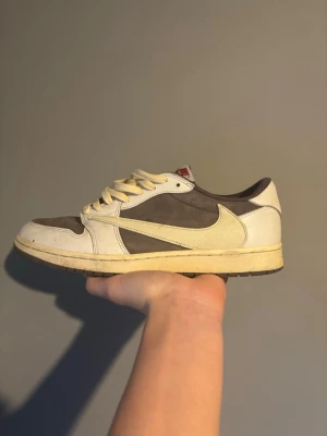 Nike Air Jordan 1 Low Travis Scott - Nike Air Jordan 1 Low Travis Scott med bruna och vita paneler, ikonisk omvänd Swoosh i beige och röda detaljer på tungan. Skon har en låg silhuett och är tillverkad i läder och mocka för en lyxig känsla. Perfekt för dig som vill sticka ut med en hypad sneaker.