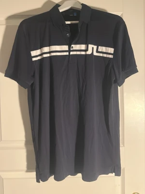 Marinblå pikétröja från J.Lindeberg - Snygg marinblå pikétröja från J.Lindeberg med vit logga och ränder över bröstet. Tröjan har korta ärmar, krage och knäppning framtill. Perfekt för en sportig och stilren look.