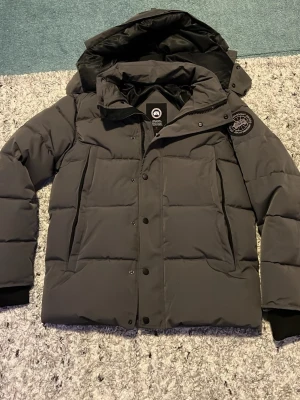 Mörkgrå pufferjacka från Canada Goose - Snygg mörkgrå pufferjacka från Canada Goose med avtagbar huva och klassisk logotyp på ärmen. Jackan har stora fickor framtill, dragkedja och tryckknappar samt ribbade muddar vid ärmsluten. Perfekt för kalla vinterdagar. Passar perfekt 170-180cm lång 👍