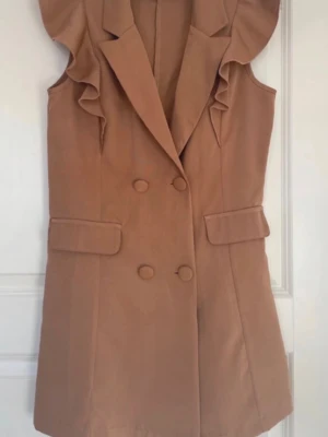 Brun väst med volangdetaljer - Snygg brun väst med dubbelknäppning och volangdetaljer vid axlarna. Västen har två fickor med lock och en klassisk krage. Perfekt för att lyfta en outfit med en trendig touch. Materialet ser ut att vara mjukt tyg, troligen polyester eller bomullsblandning.
