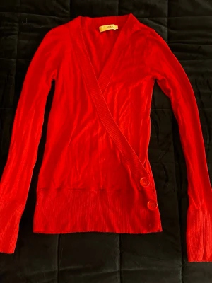 Röd omlottkofta från Zoul - Snygg röd omlottkofta från Zoul med långa ärmar och två dekorativa knappar framtill. Koftan har ribbade muddar vid ärmslut och nederkant samt en djup v-ringning. Perfekt för att addera färg till din outfit.