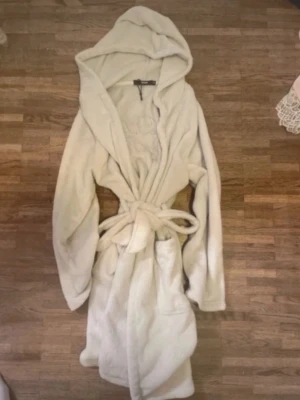 Beige fluffig morgonrock med huva - Mysig beige morgonrock i fluffigt material med stor huva och knytskärp i midjan. Två stora fickor framtill och långa ärmar. Perfekt för att chilla hemma och hålla sig varm under kalla morgnar.