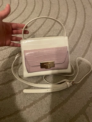 Vit och ljusrosa handväska med krokoprägling - Snygg handväska i vitt och ljusrosa med krokodilpräglat mönster. Väskan har både handtag och justerbar axelrem samt ett guldfärgat lås framtill. Perfekt accessoar för att lyfta din outfit.