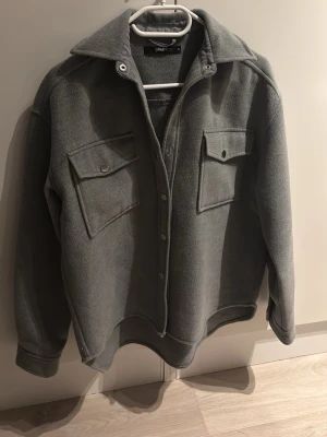 Grå overshirt från Gina Tricot - Stilren grå overshirt från Gina Tricot i mjukt fleece-liknande material. Två stora bröstfickor med knappstängning, klassisk krage och tryckknappar framtill. Perfekt lager-på-lager-plagg med avslappnad passform och långa ärmar.