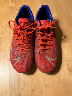Nike Mercurial röda fotbollsskor - Säljer ett par Nike Mercurial fotbollsskor i rött med blå och gröna detaljer. Skorna har snörning, låg profil och mönstrad ovansida för bättre bollkontroll. Yttersulan har fasta dobbar och insidan är blå. Perfekta för dig som vill ha snabbhet och stil på planen.