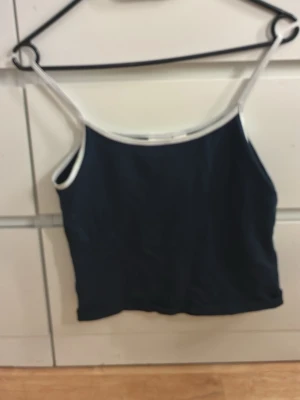 Mörkblå linnetopp med vita kanter - Enkel och stilren mörkblå linnetopp med vita kanter runt hals och axelband. Perfekt att matcha med jeans eller kjol för en avslappnad look.