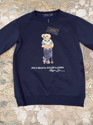 Mörkblå sweatshirt Polo Bear Ralph Lauren - Mörkblå sweatshirt från Polo Ralph Lauren med ikoniska Polo Bear-trycket framtill. Tröjan har rund halsringning, ribbade muddar och är tillverkad i mjukt bomullsmaterial. Perfekt för en avslappnad och stilren look.