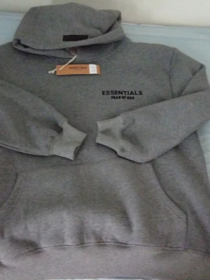 Grå Essentials Fear of God hoodie - Snygg grå hoodie från Essentials Fear of God med svart logga på bröstet. Klassisk modell med huva och stor magficka framtill. Perfekt för en avslappnad streetwear-look. Mjuk och skön bomullsblandning.