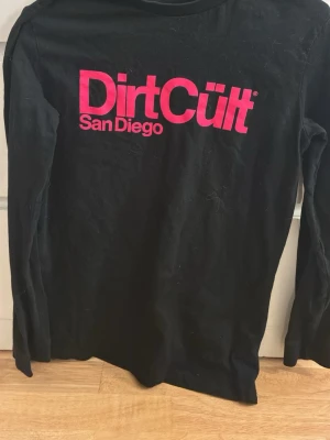 Svart långärmad DirtCült t-shirt - Svart långärmad t-shirt från DirtCült med stor rosa logga och texten 'San Diego' på bröstet. Skön passform och perfekt för dig som gillar streetwear. Materialet känns mjukt och passar till många olika stilar.