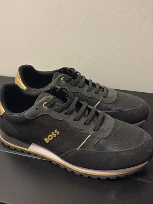 Svarta BOSS sneakers med gulddetaljer - Snygga svarta sneakers från BOSS med gulddetaljer och logga på sidan. Skorna har lågt skaft, svarta skosnören och en vit och guldig rand på sulan. Ovandelen är i textil och syntet, perfekt för en stilren look.