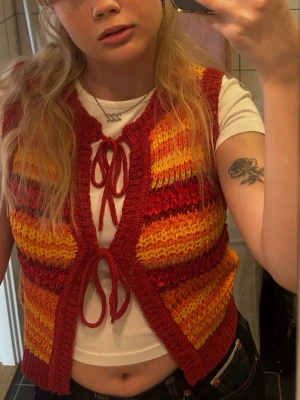 Randig stickad väst i rött och orange - Säljer en färgglad stickad väst med ränder i incident rött, orange och gult. Västen har två knytband framtill och ribbade kanter. Perfekt för att addera lite retrovibbar till din outfit.
