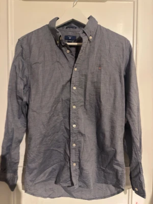 Blå skjorta från Gant med knappar - Klassisk blå skjorta från Gant med button-down krage och vita knappar framtill. Skjortan har lång ärm och en liten broderad logga på bröstet. Tillverkad i bomull för en bekväm känsla och stilren look.